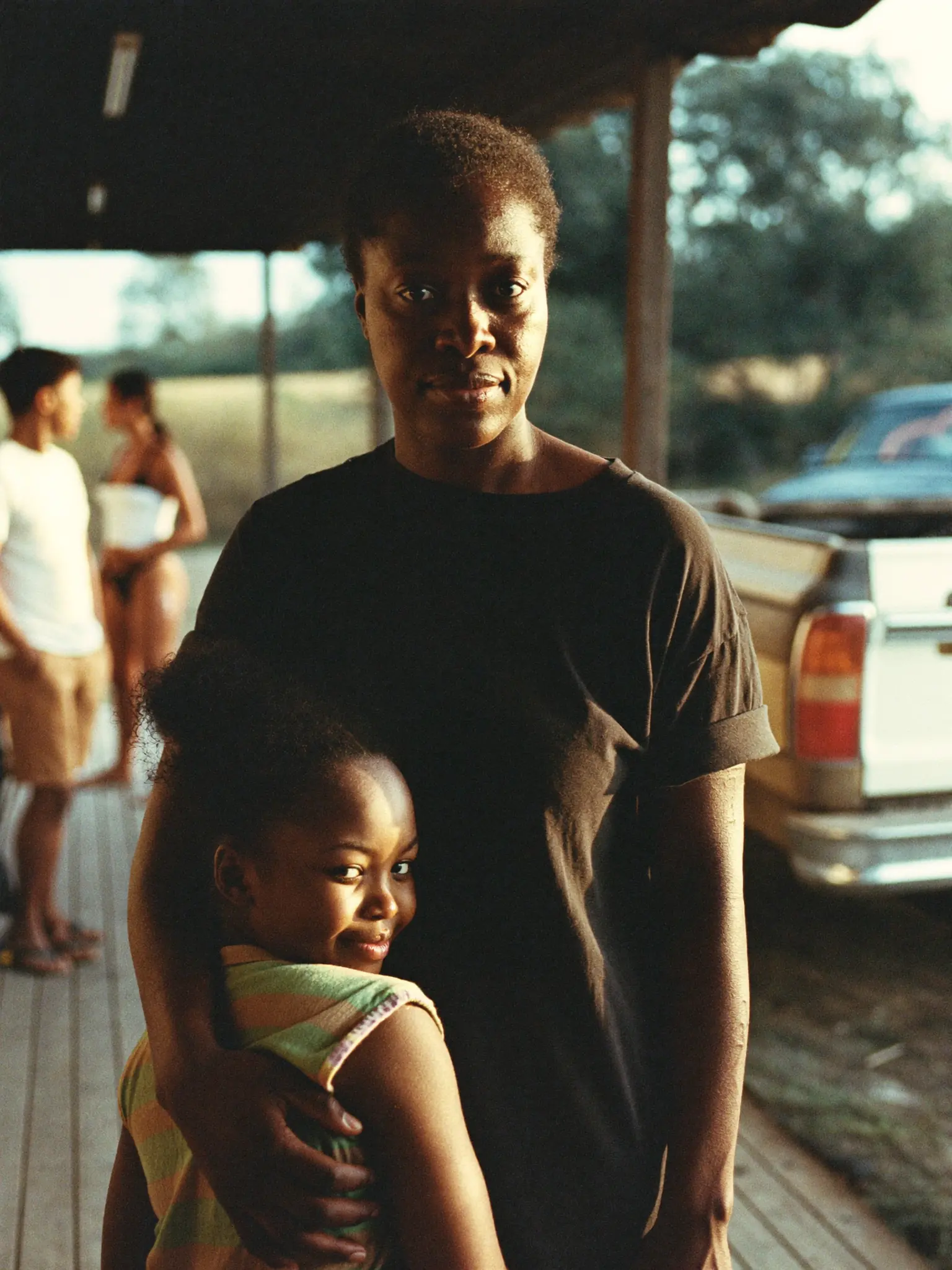 A mom in a black tee with her daughter held close on a porch at golden hour.